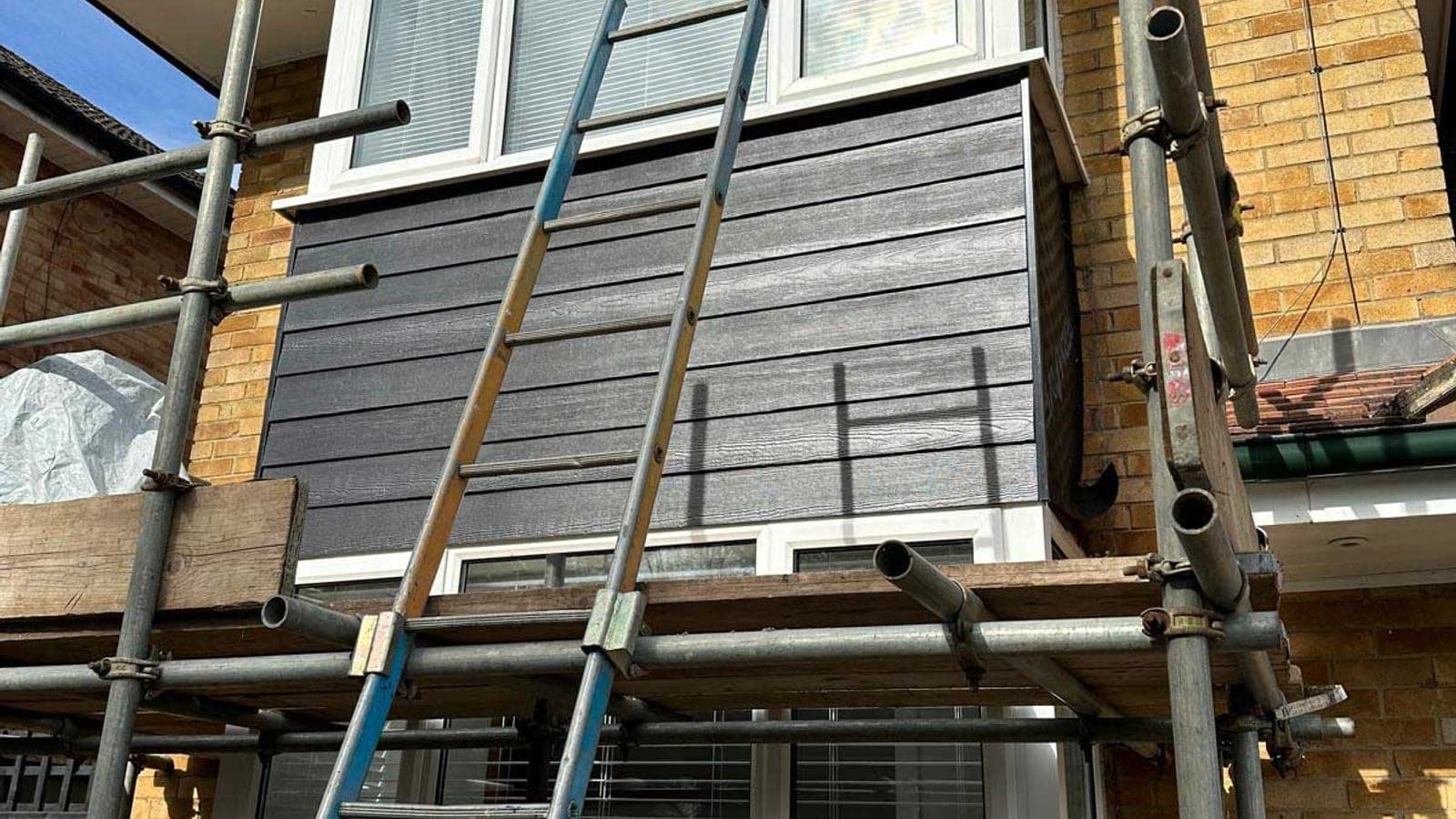 Dark grey weatherboard cladding half fitted to a first-floor bay, with ladders and scaffold in front