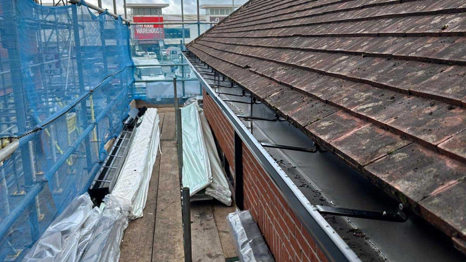 Run of new gutter brackets fixed along the eaves of a weathered clay tiled roof, seen from the scaffold boards