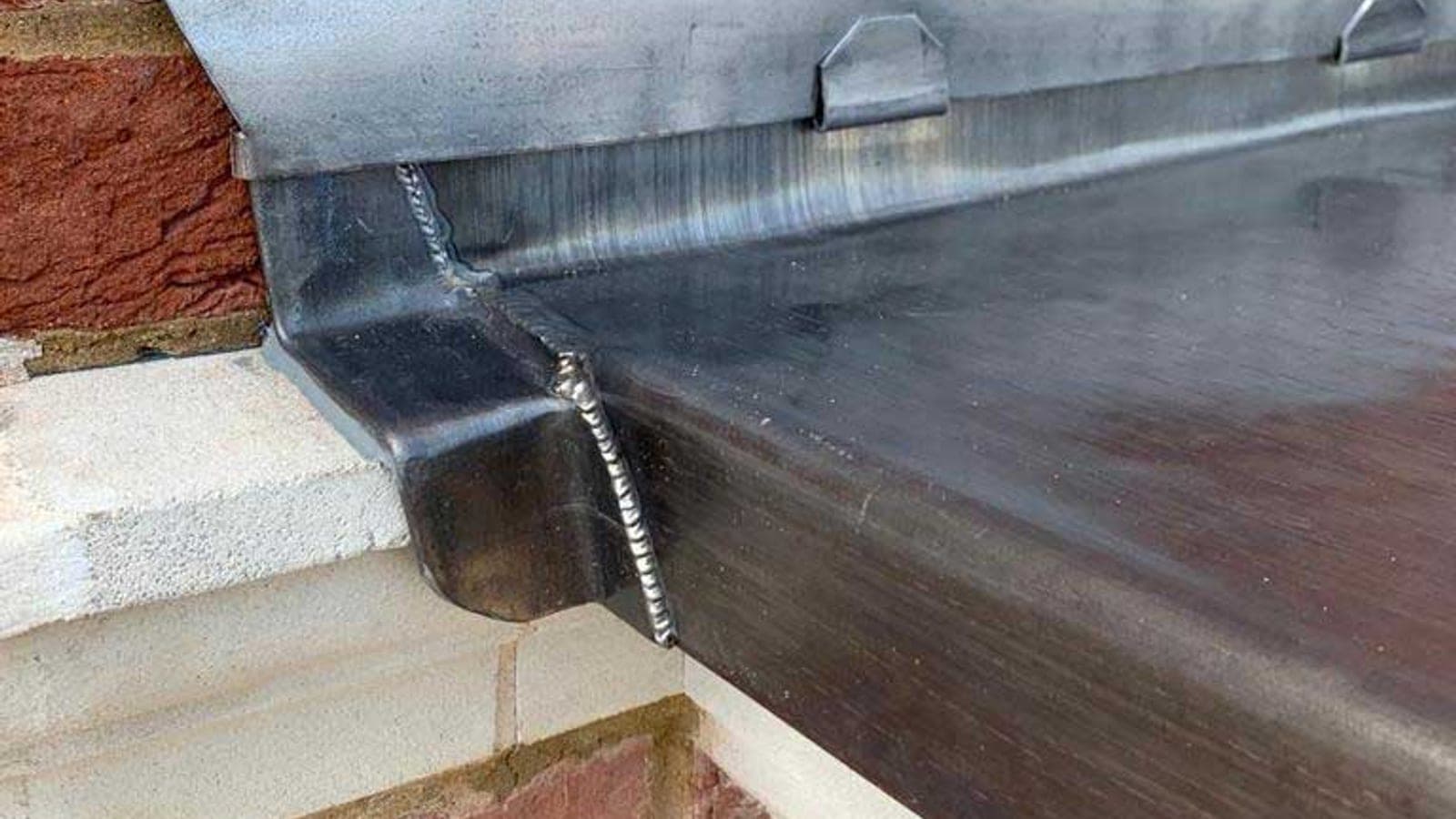 Close-up of a lead-lined parapet gutter with a welded corner joint against red brickwork