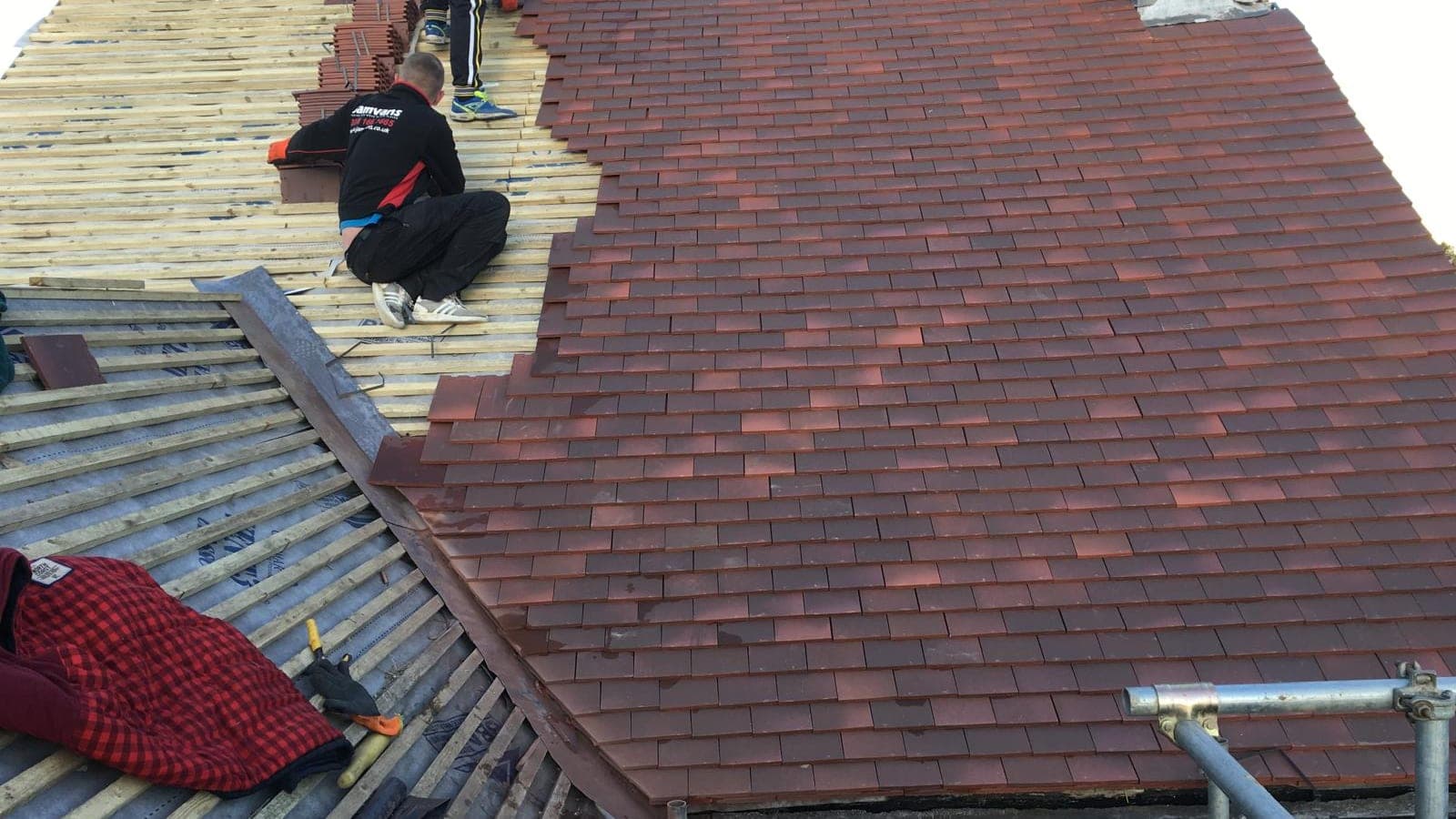 New red clay plain tiles being laid over fresh battens beside a lead valley, two roofers working up the slope