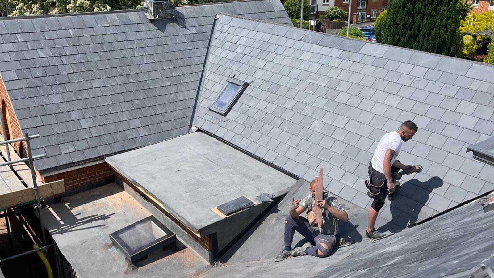 Two roofers laying slate on a pitched roof beside a finished lead flat roof with rooflights