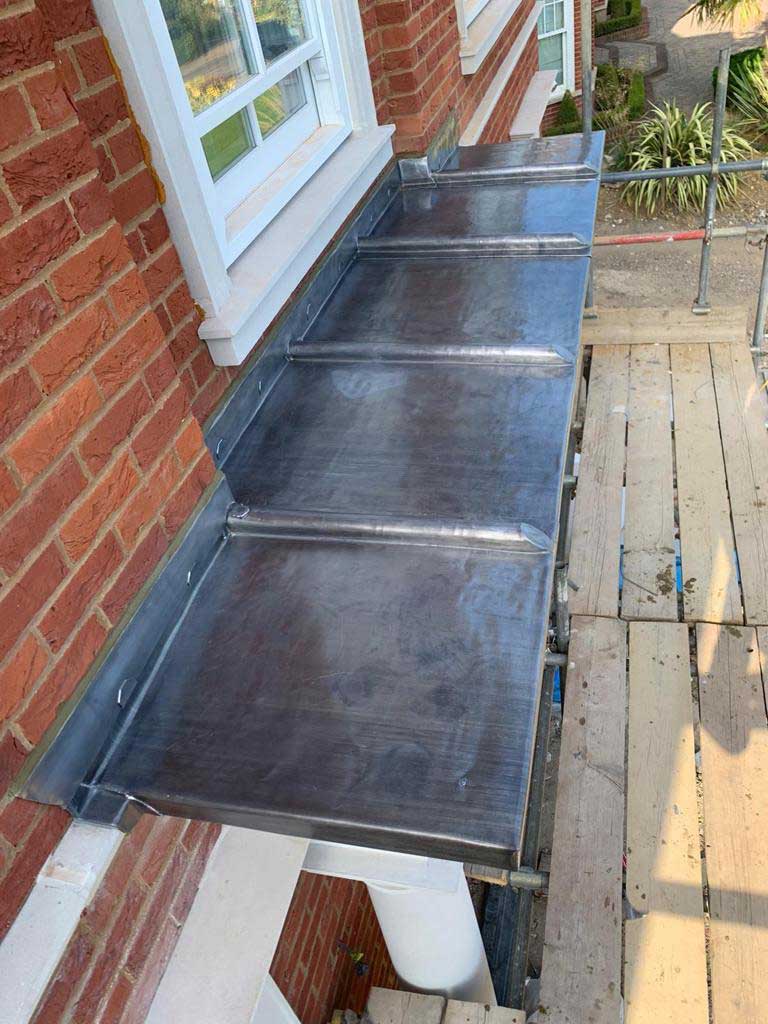 Lead-covered bay roof with welded rolls running down to a drip, above a painted stone cornice